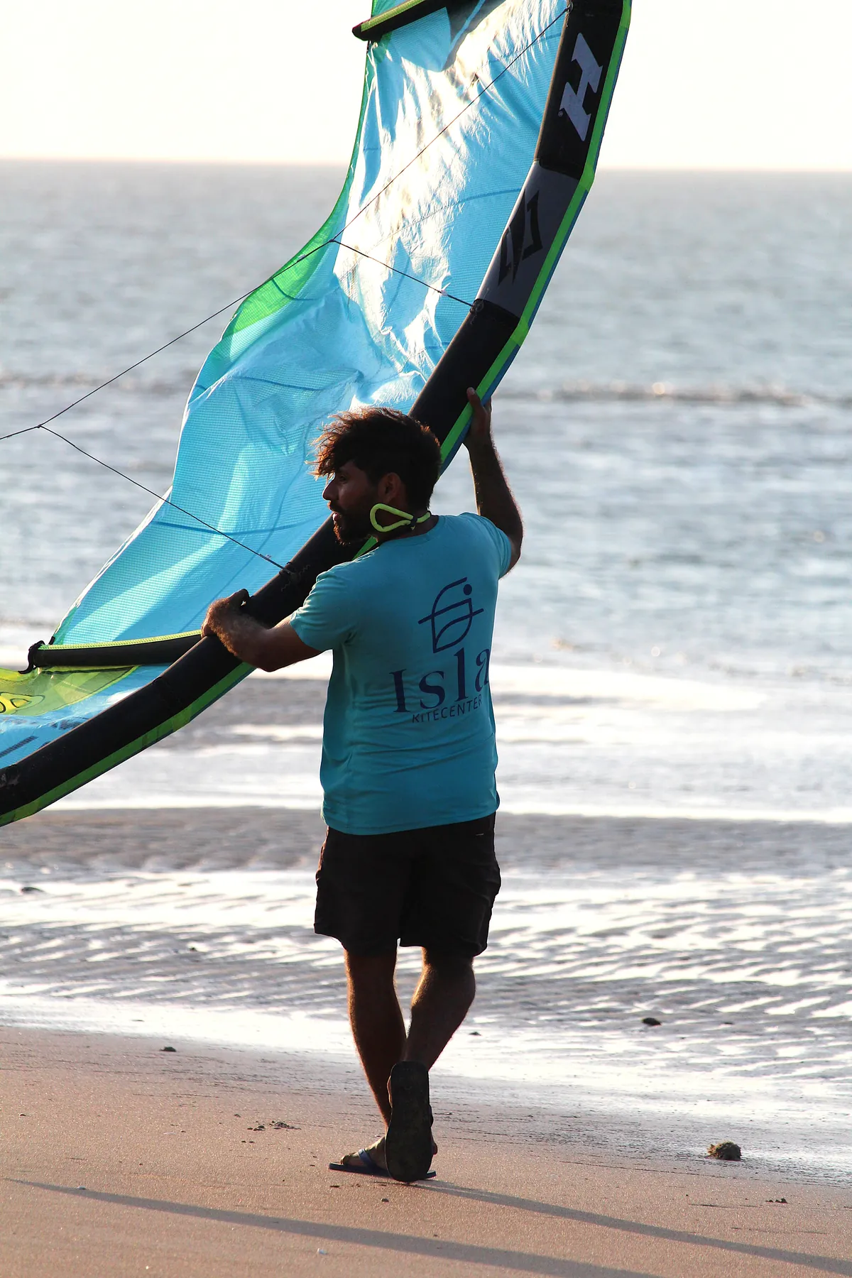 Instrutor com camiseta Isla Kite Center segurando pipa Naish azul-turquesa na praia da Ilha do Guajirú ao entardecer dourado